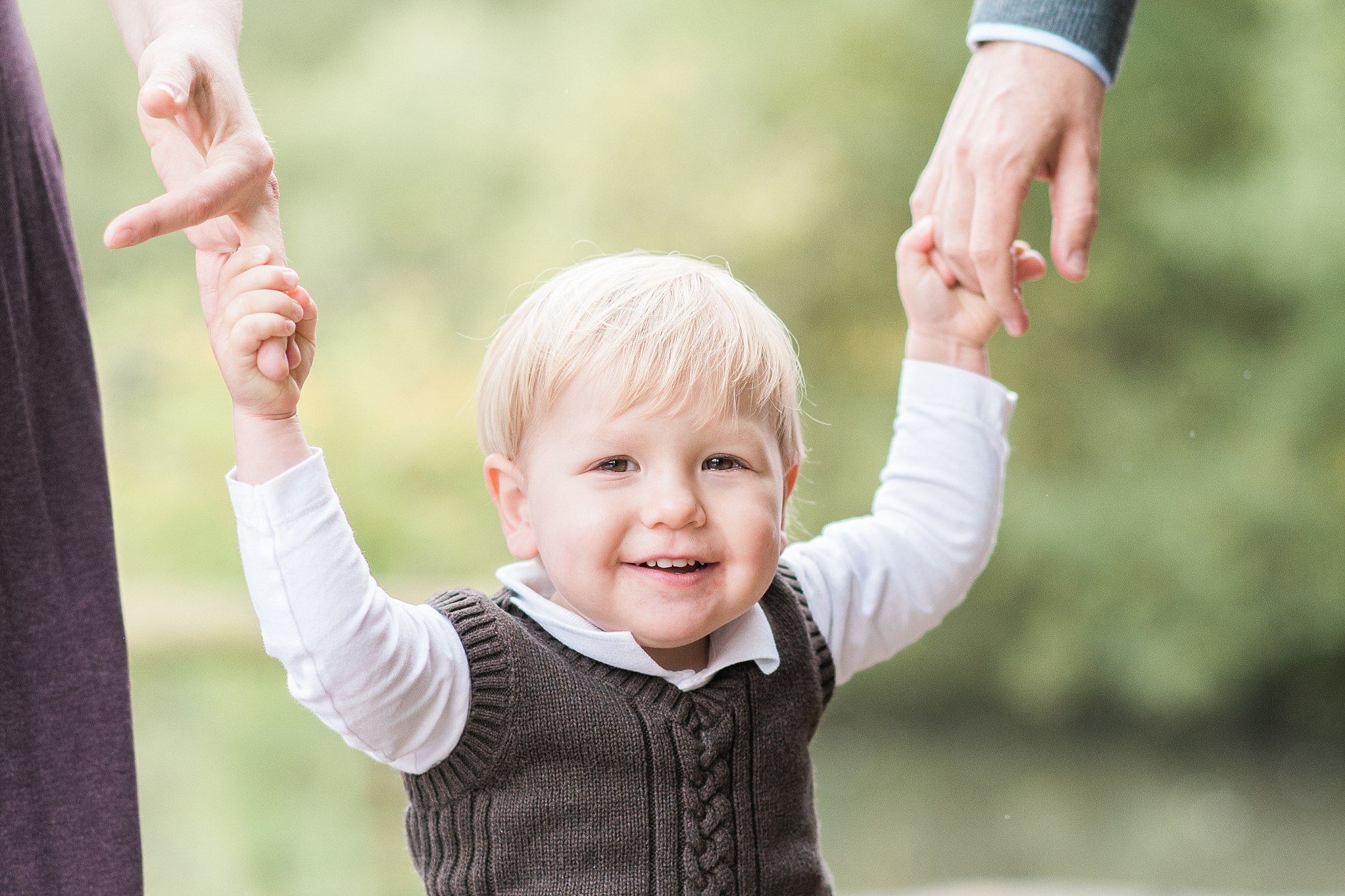Blonde toddler walking with parents' support during an outdoor family photo session in Leeds, Yorkshire